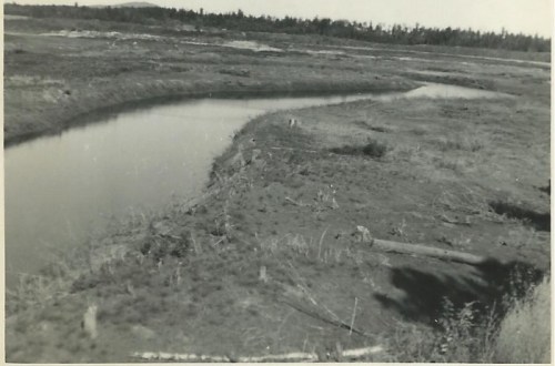A winding river flows through a barren landscape, with sparse vegetation and distant trees in the background.
