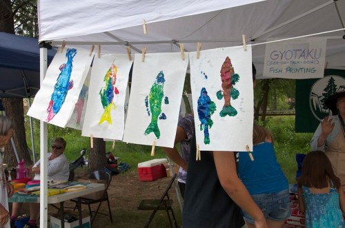 Colorful fish prints hanging on a clothesline under a tent at an outdoor event, with people in the background.