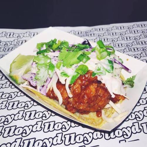 A delicious taco topped with fried chicken, slaw, cilantro, and lime, served in a paper tray on a patterned background.