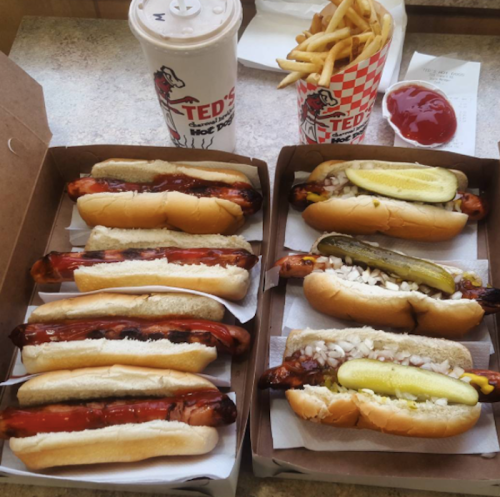 A takeout box with various hot dogs, fries, a drink, and ketchup on a table.