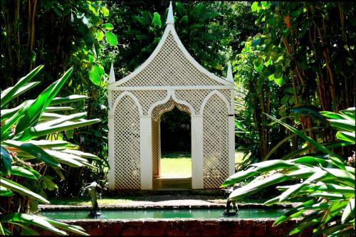 A white lattice gazebo surrounded by lush greenery and a small pond with statues in a tranquil garden setting.