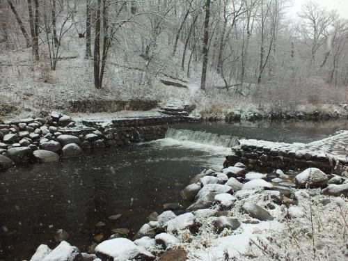 A snowy landscape featuring a river with a small waterfall and rocky banks, surrounded by trees.