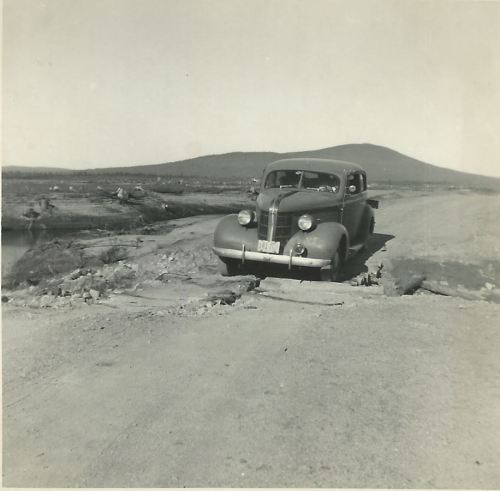 A vintage car parked on a dirt road with a mountainous landscape in the background.