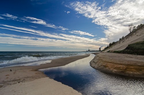 A serene beach scene with gentle waves, a winding stream, and a cloudy sky in the background.