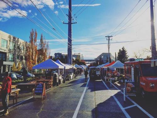 A vibrant street market with colorful tents, food trucks, and clear blue skies, lined with trees and power lines.