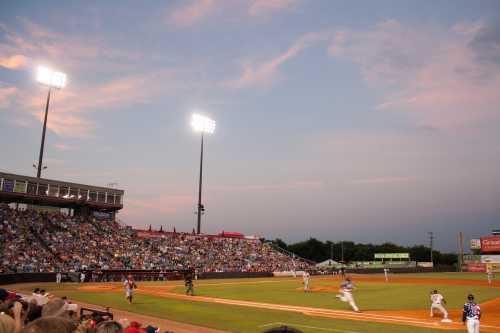 A baseball game in progress at dusk, with a crowd in the stands and bright stadium lights illuminating the field.