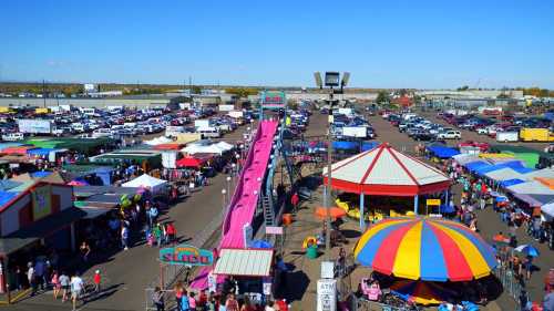 A vibrant outdoor fair with colorful tents, a pink slide, and crowds of people enjoying the festivities under a clear blue sky.