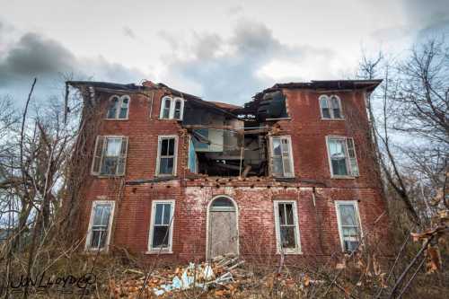 A dilapidated brick house with a collapsed roof, surrounded by overgrown vegetation and a cloudy sky.