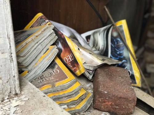 A pile of old magazines and a brick, resting on a dusty surface near a wooden structure.