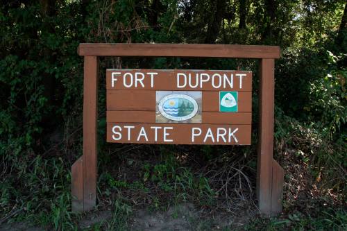 Wooden sign for Fort Dupont State Park, featuring park logos and surrounded by greenery.