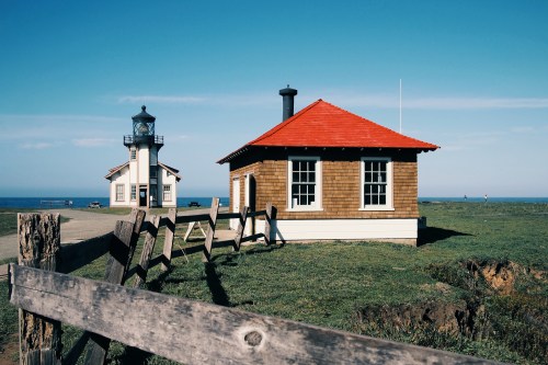 A coastal scene featuring a lighthouse and a wooden building with a red roof, surrounded by green grass and a blue sky.