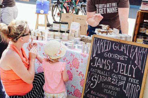 A woman and a child examine jars at a market stall featuring gourmet jams and jellies. A sign lists flavors available.