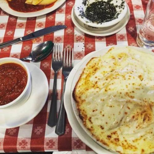 A plate of cheesy baked pasta with marinara sauce, accompanied by a small bowl of pesto and utensils on a checkered tablecloth.