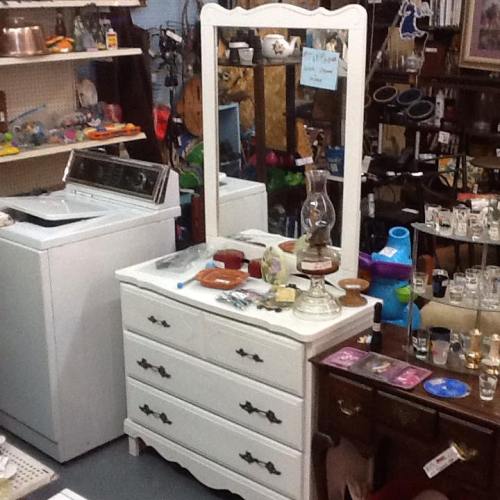 A vintage dresser with a mirror, a washing machine, and various glass items in a cluttered thrift store setting.