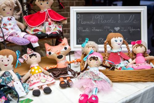 A display of handmade dolls in various outfits, including a fox doll, arranged on a table with a chalkboard sign.