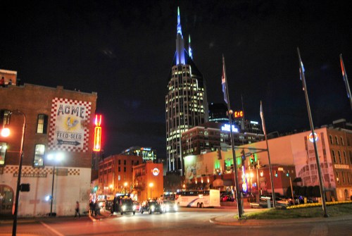 A city street at night featuring a tall, illuminated skyscraper and vibrant neon signs.