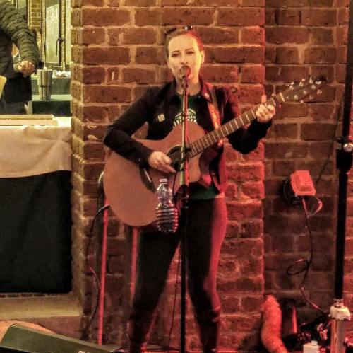 A woman sings and plays guitar in a cozy venue, with a brick wall backdrop and a person visible in the background.