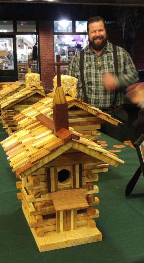 A smiling man stands beside a detailed wooden model of a log cabin, showcasing craftsmanship at a display table.