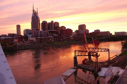 Sunset over Nashville skyline with vibrant colors reflecting on the river and a sculpture in the foreground.