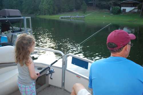 A child and an adult fishing from a boat on a calm lake surrounded by trees.