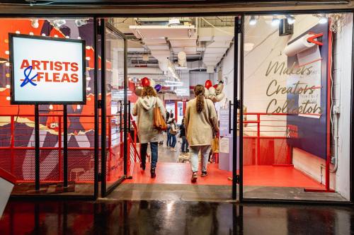Two people walk through a vibrant entrance labeled "Artists & Fleas," leading into a creative marketplace.