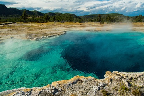 A vibrant blue hot spring surrounded by rocky terrain and lush greenery under a cloudy sky.