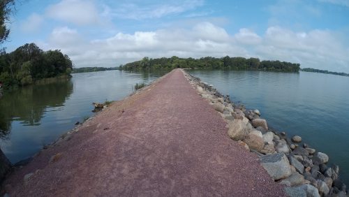 A wide path made of gravel stretches between calm waters, flanked by trees under a partly cloudy sky.