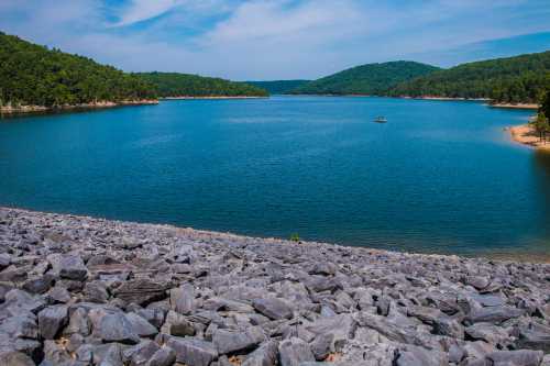 A serene lake surrounded by green hills, with a rocky shoreline and a clear blue sky above.