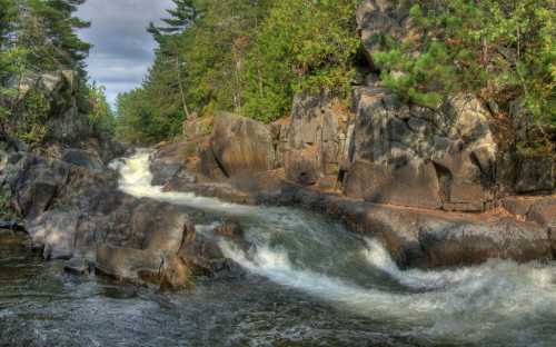 A serene river flows through rocky terrain, surrounded by lush greenery and trees under a cloudy sky.