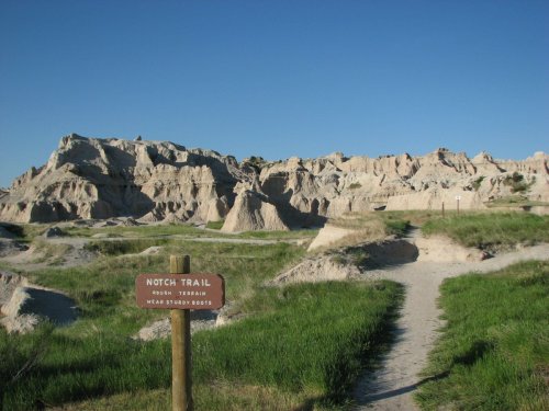 A trail sign for Notch Trail in a grassy area, with rugged rock formations and a clear blue sky in the background.