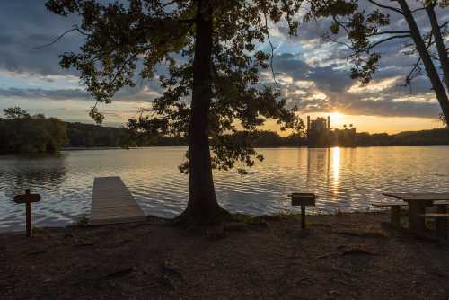 A serene lake at sunset, with a wooden dock, trees, and a distant building silhouetted against the colorful sky.