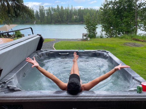 A person relaxes in a hot tub with arms outstretched, surrounded by greenery and a river in the background.