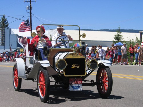A vintage Ford car in a parade, with two people waving, surrounded by spectators and American flags.