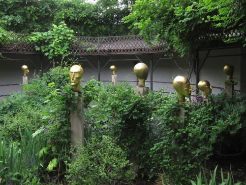 A garden with lush greenery featuring several golden head sculptures on stone pedestals.