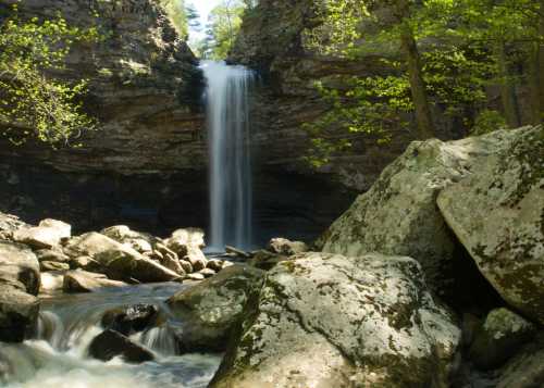 A serene waterfall cascades down rocky cliffs, surrounded by lush greenery and smooth stones in a tranquil forest setting.