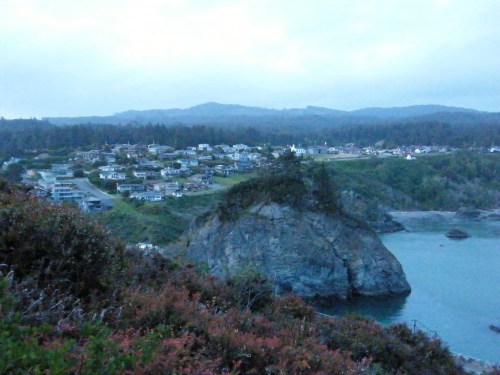 A coastal view of a small town nestled among hills, with rocky shores and a calm sea under a cloudy sky.