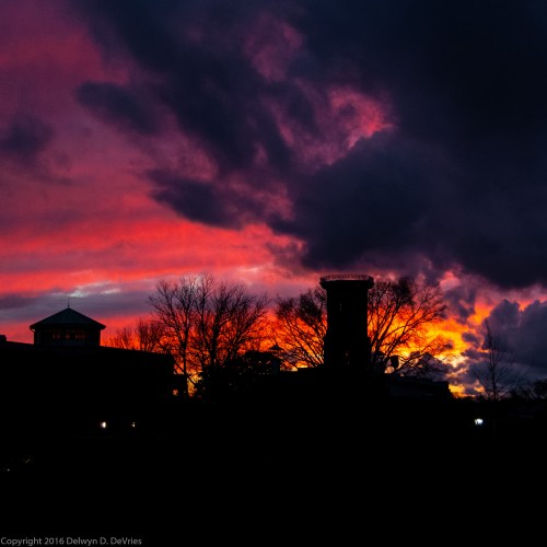 A vibrant sunset with deep orange and purple hues, silhouetting trees and buildings against the colorful sky.