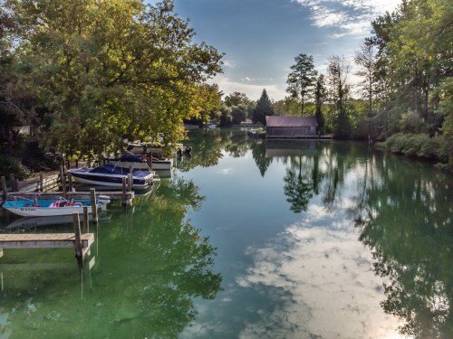 A serene river scene with boats docked, surrounded by trees and reflecting clouds in the calm water.