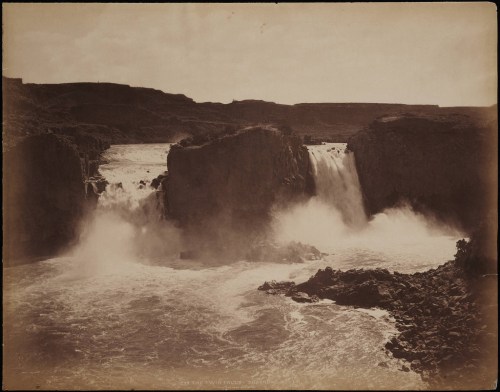 A historic black-and-white photo of a waterfall cascading over rocky cliffs, surrounded by mist and rugged terrain.