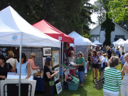 A bustling outdoor market with vendors under white and red tents, shoppers browsing art and crafts in a green park setting.