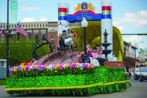A colorful parade float decorated with flowers, featuring children and a backdrop of festive decorations.