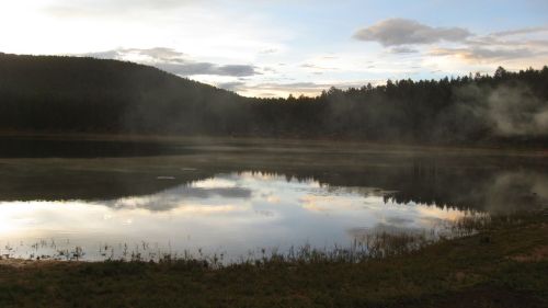 A serene lake reflects the sky and surrounding hills, with mist rising from the water at dawn.