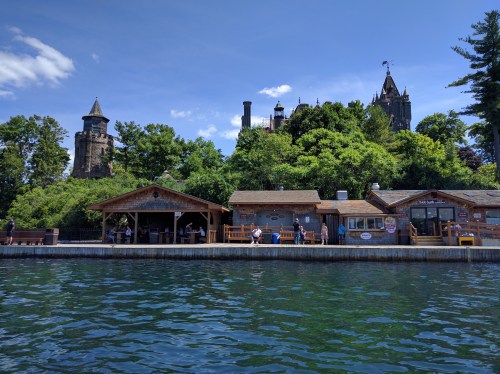 A scenic view of a lakeside building and a castle on a hill, surrounded by lush greenery under a blue sky.