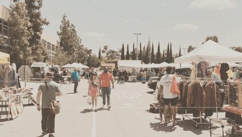A bustling outdoor market with tents, vendors, and shoppers walking along a paved path under a sunny sky.
