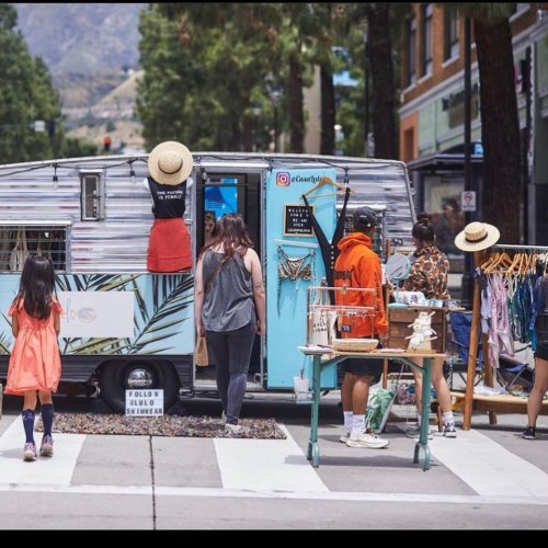 A vibrant outdoor market scene with a vintage trailer, clothing racks, and people browsing in a tree-lined street.