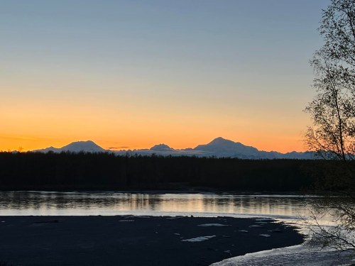 Sunset over mountains reflected in a calm river, with a silhouette of trees in the foreground.