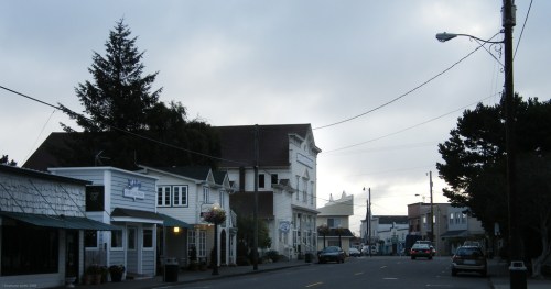 A quiet street scene featuring charming buildings, trees, and overcast skies. Cars are parked along the road.