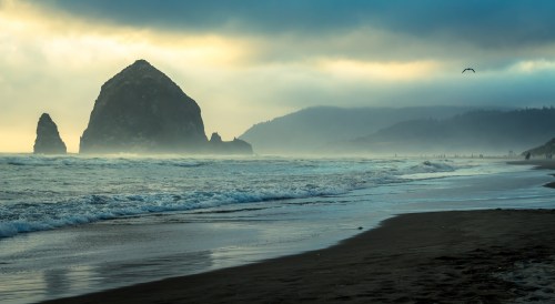 A misty beach scene with large rock formations, gentle waves, and a cloudy sky in the background.