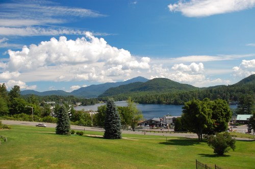 A scenic view of a lake surrounded by mountains, with green grass and trees in the foreground under a blue sky.