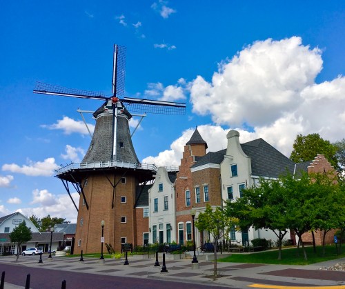 A large windmill next to a charming building under a blue sky with fluffy clouds. Green trees line the walkway.